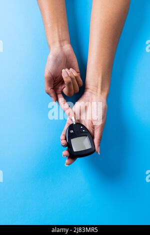 Beschnittene Hände einer afroamerikanischen mittleren erwachsenen Frau, die Zucker mit Glucometer auf blauem Hintergrund untersucht Stockfoto