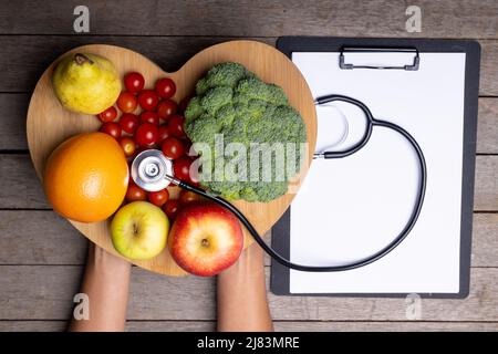 afroamerikanische Frau mit Herzform, Gemüse, Obst, Stethoskop nach Bericht auf dem Tisch Stockfoto