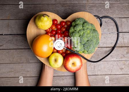 afroamerikanische Frau mit mittlerem Erwachsenen, die eine Herzform mit Gemüse, Obst und Stethoskop auf dem Tisch hält Stockfoto