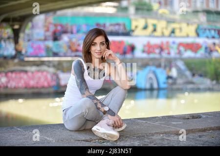 In einer Stadt am Ufer eines Flusses sitzt eine junge, junge Frau langen braunen Haaren und Tattoo. Sie traegt eine Freizeitregelung mit Sportschuhen Stockfoto
