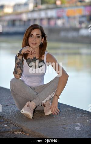 In einer Stadt am Ufer eines Flusses sitzt eine junge, junge Frau langen braunen Haaren und Tattoo. Sie traegt eine Freizeitregelung mit Sportschuhen Stockfoto