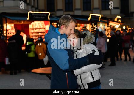 Vor den beleuchteten Verkaufsstaenden an einem Weihnachtsmarkt steht ein junges verliebtes Paar und hat sich im Arm Stockfoto