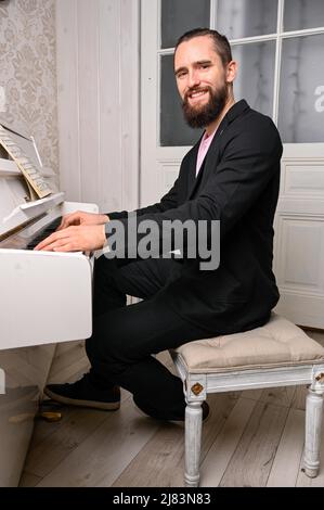 An einem Klavier sitzt ein junger Mann mit Vollbart und kurzen Haaren. Er blicks to camera waehrend er beside haende on the tasten liegen has. Der Stockfoto