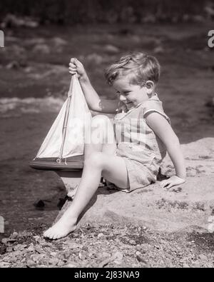 1930S LÄCHELNDER BARFUSS-JUNGE IN EINEM SEERSUCKER-SPIELANZUG, DER AUF EINEM FELSEN NEBEN DEM STROM SITZT, UM SPIELZEUG ZU STARTEN SEGELBOOT - J6770 HAR001 HARS HALBLANGER BACH MÄNNCHEN SEGELSTART ÜBER SEGELBOOT B&W SOMMERGLÜCK FRÖHLICHES HOCHWINKEL-ABENTEUER AUFREGUNG DURCH VON AUF ZU BARFUSS LÄCHELT SPIELEN ANZUG KONZEPTIONELL FRÖHLICH SEGEL BOOT BABY JUNGE WACHSTUM JUGENDLICHE SAISON SEERSUCKER SCHWARZ UND WEISSE KAUKASISCHE ETHNIZITÄT CREEK HAR001 ALTMODISCH Stockfoto