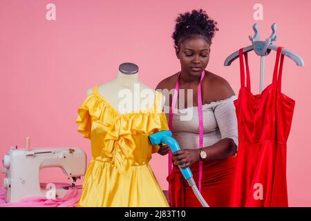 afroamerikanische Schneiderin hält Dampfdampfgerät Heißdampfgenerator für Seide rot und gelb Kleid im Trockenreinigungssalon, Studio rosa Stockfoto