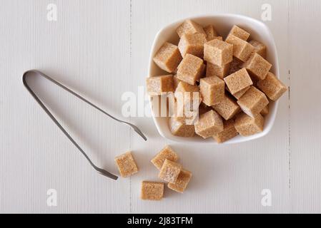 Würfelförmiger Rohrzucker in Holzschüssel auf einem weißen Holztisch mit silberner Zuckerzange. Flach liegend, Blick von oben nach unten, keine Leute. Stockfoto