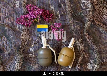 Eine militärische Handgranate liegt auf einem Tarnnetz und neben der Flagge der Ukraine und einem Zweig von Flieder im Frühjahr der Krieg in der Ukraine, der Krieg in Großbritannien Stockfoto