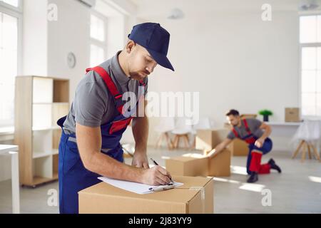 Professioneller männlicher Mover in Overalls macht Notizen auf der Zwischenablage, während er in der Nähe von Kartons steht. Stockfoto