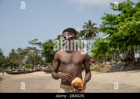 Ein junger schwarzer Mann mit einer Kokosnuss in den Händen, der im Urlaub eine Sonnenbrille trägt und Spaß hat. Stockfoto