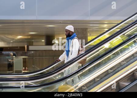 Afroamerikanischer Passagiermann mit Koffer steht auf Rolltreppe, hält Handlauf im Flughafenterminal Stockfoto