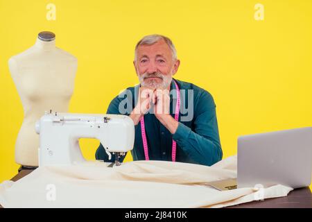 Angenehmer alter Mann, der in Schneideratelier arbeitet und Kleidung an der Nähmaschine macht, gelber Hintergrund Stockfoto