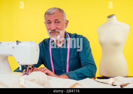 Angenehmer alter Mann, der in Schneideratelier arbeitet und Kleidung an der Nähmaschine macht, gelber Hintergrund Stockfoto