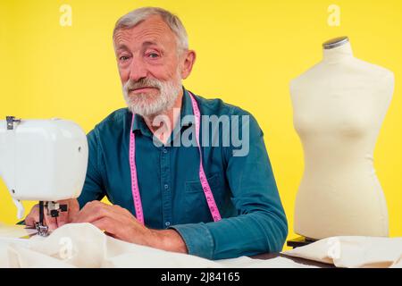 Angenehmer alter Mann, der in Schneideratelier arbeitet und Kleidung an der Nähmaschine macht, gelber Hintergrund Stockfoto