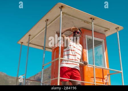 afroamerikanischer junger Mann mit Sonnenbrille, der die Augen auf der Rettungsschwimmerhütte am Strand gegen den Himmel schützt Stockfoto