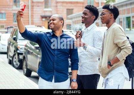 Eine Gruppe von drei modischen coolen afroamerikanischen Jungs Studenten auf der Straße kommunizieren Blick auf Smartphone nehmen Fotos Selfie auf Telefon in Stockfoto