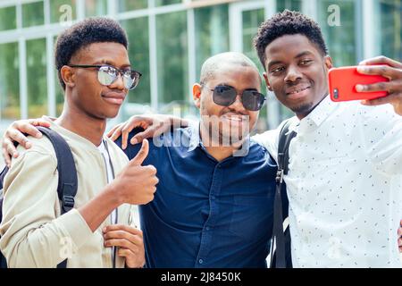 Eine Gruppe von drei modischen coolen afroamerikanischen Jungs Studenten auf der Straße kommunizieren Blick auf Smartphone nehmen Fotos Selfie auf Telefon in Stockfoto