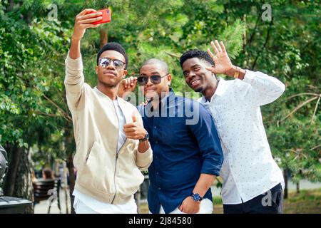 Eine Gruppe von drei modischen coolen afroamerikanischen Jungs Studenten auf der Straße kommunizieren Blick auf Smartphone nehmen Fotos Selfie auf Telefon in Stockfoto