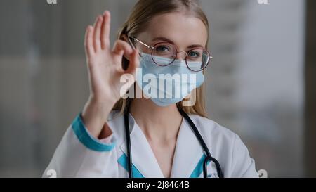 Porträt kaukasischen Arzt Spezialist Therapeut Krankenschwester Chirurg Frau Mädchen in einer Brille in medizinischen Schutz Gesichtsmaske Blick auf die Kamera zeigt ok Stockfoto