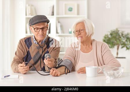 Älterer Ehemann, der seinen Blutdruck bei seiner Frau überprüft und zu Hause an einem Tisch sitzt Stockfoto