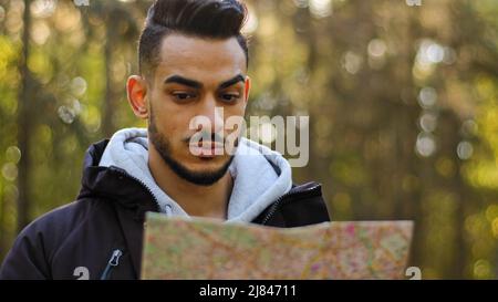 Männliches Portrait Nahaufnahme junger indischer männlicher Tourist, der im Wald im Freien steht, schaut auf die Papierkarte, die neue bequeme Navigationsroute legt Stockfoto