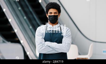 afroamerikanischer Mann Kerl Restaurant Café männlich Arbeiter in medizinische Maske aussehende Kamera posiert über Arme Kellner Verkäufer trägt Schürze stehen an Stockfoto