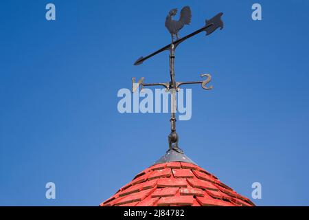 Wettervane in Form eines Hahns auf einem kegelförmigen Dach aus Zedernholz. Stockfoto