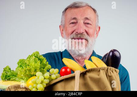 Alter älterer Großvater hält wiederverwendbare Einkaufstasche aus Baumwolle voller natürlichem Bauerngemüse: Mais, Bananen, Auberginen und Trauben in Studioweiß Stockfoto