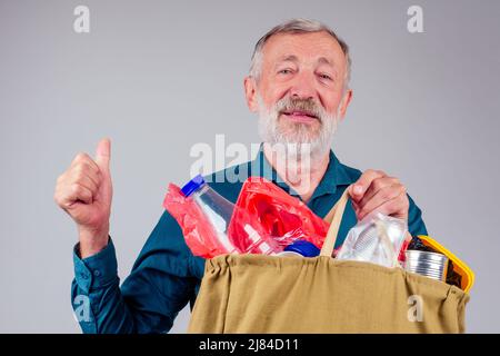 Alter Großvater hält Baumwollektasche voller Müll auf weißem Hintergrund im Studio Stockfoto