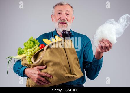 Alter älterer Großvater, der sich im Studio zwischen Einweg-Polyethylen-Paket und wiederverwendbarer Einkaufstasche aus Baumwolle mit natürlichem Bauerngemüse entschied Stockfoto