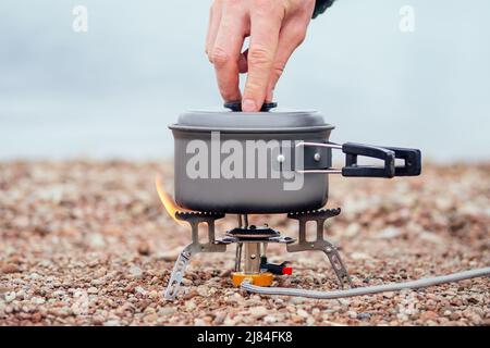 Die Pfanne mit dem Brei steht auf dem Gasbrenner (Campingkocher). Auf dem Hintergrund des Flusses Stockfoto