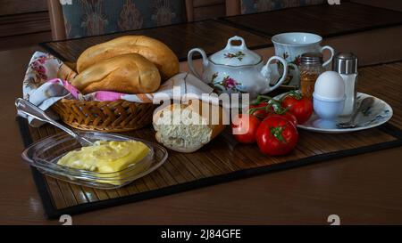 Ein weich gekochtes Ei zum Frühstück mit frischen Brötchen, Butter, einer Tasse Tee und einem Haufen schöner Tomaten. Das Foto kann auf Leinwand und Poster verwendet werden. Stockfoto