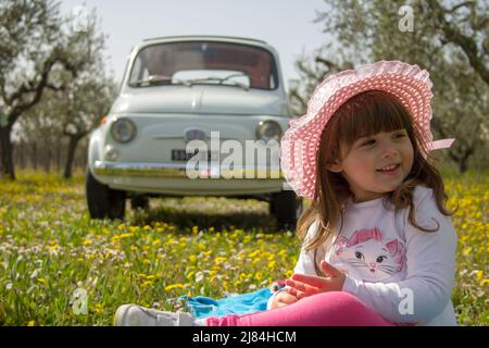Foto eines entzückenden lächelnden kleinen Mädchens in einem Hut, das auf einer blühenden Wiese mit einem Oldtimer im Hintergrund während einer Reise nach Italien sitzt Stockfoto