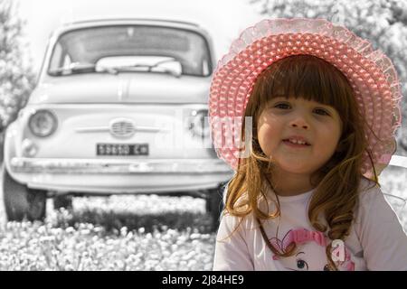 Foto eines entzückenden lächelnden kleinen Mädchens in einem Hut, das auf einer blühenden Wiese mit einem Oldtimer im Hintergrund während einer Reise nach Italien sitzt Stockfoto