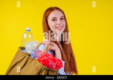 Schöne rothaarige Ginder glückliche Frau hält Baumwolle Öko-Beutel mit Sortierung Papierkorb: Plastikflasche, Einweg-Paket im Studio gelben Hintergrund Stockfoto