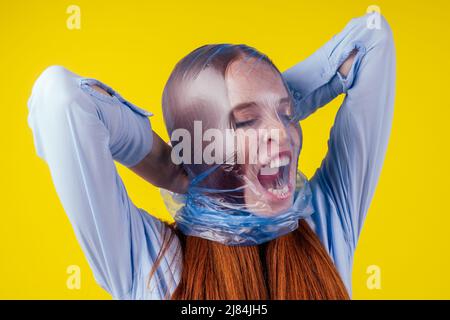 Rothaarige Ingwer europäische Frau Erstickungskopf in blauen Polyethylen-Paket in Studio gelben Hintergrund. Eco Verschmutzung Cellophan Film Konzept Stockfoto