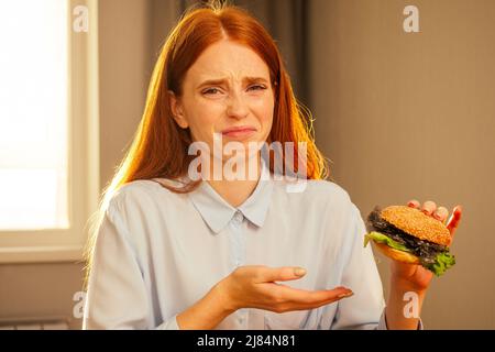 Rothaarige Ingwer Mädchen essen gefälschte Burger mit Brötchen Sesam und Kunststoff Zellophan Film Müll, Trinkwasser aus Einweg-Tasse mit nicht-Öko-Schlauch an Stockfoto