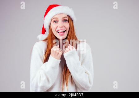 Schöne rothaarige Ingwer Frau trägt weißen Strickpullover und weihnachtsmann Hut Finger Kreuz macht Wünsche im Studio Hintergrund Stockfoto