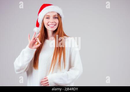 Schöne rothaarige Ingwer Frau trägt weißen Strickpullover und weihnachtsmann Hut im Studio Hintergrund Stockfoto