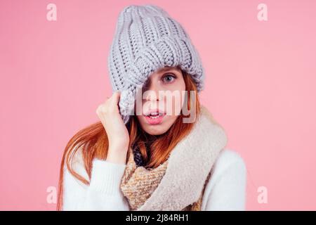Schüchterne Rothaarige Frau, die einen gestrickten spitzen Hut auf den Kopf setzt, Auge und offenen Mund in Studio rosa Hintergrund versteckt Stockfoto