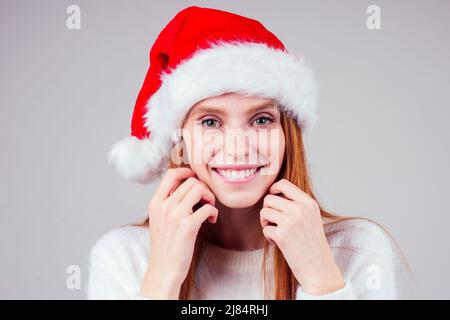 Schöne rothaarige Ingwer Frau trägt weißen Strickpullover und weihnachtsmann Hut erwartet Magie im Studio Hintergrund Stockfoto