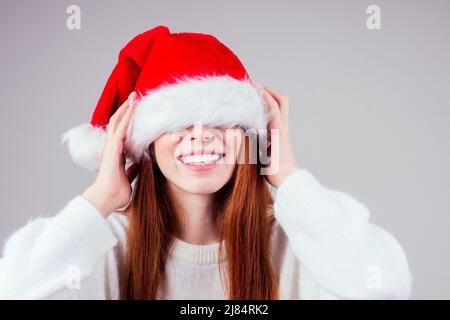 Schüchtern schöne rothaarige Ingwer Frau trägt weißen Strickpullover und versteckt Auge in santa claus Hut Studio Hintergrund Stockfoto