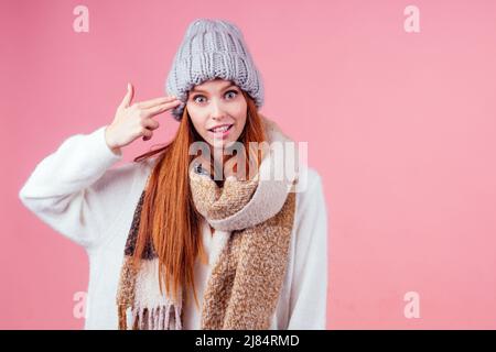 Rothaarige Ingwer Frau in Hut, gestrickten Pullover und Schal zeigen Finger Hände wie Waffen Geste auf den Kopf in Studio rosa Hintergrund copyspase Stockfoto