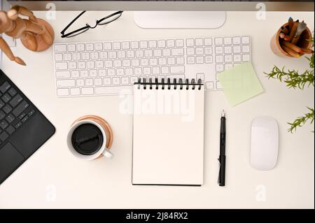 Moderner weißer Schreibtisch mit leerem spiralförmigem Notizblock, Kaffeetasse, Tastatur und Schreibwaren. Draufsicht Stockfoto