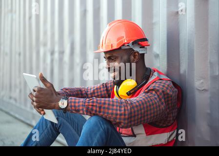 Nahaufnahme glücklich lächelnder afroamerikanischer Ingenieur mit Schutzweste und Helm, sitzend und mit Mobiltelefon bei logistischen Frachtgut contai Stockfoto