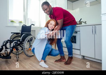 Junge bärtige, behinderte rothaarige Ingwerfrau im Rollstuhl und ihr afroamerikanischer Ehemann in Kichen Stockfoto