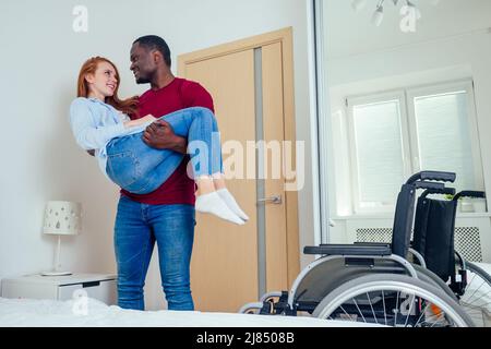 Junge bärtige, behinderte rothaarige Ingwerfrau und ihr afroamerikanischer Ehemann im Schlafzimmer. Mann hält sie an den Händen und hilft, ein Bett zu erreichen Stockfoto