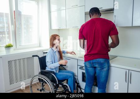 Junge bärtige, behinderte rothaarige Ingwerfrau im Rollstuhl und ihr afroamerikanischer Ehemann in Kichen Stockfoto