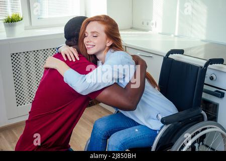 Junge bärtige, behinderte rothaarige Ingwerfrau im Rollstuhl und ihr afroamerikanischer Ehemann in Kichen Stockfoto