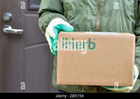 Logo iHerb auf Karton in Kurierhänden. IHerb ist ein Unternehmen für pflanzliche Produkte, bietet und liefert Gesundheits- und Wellness-Produkte Stockfoto