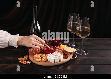 Die Hand der Frau nimmt ein Stück Käse aus dem Vorspeisen-Brett mit verschiedenen Käsesorten, Fleisch, Wurstrosette, Trauben und Keksen. Stockfoto